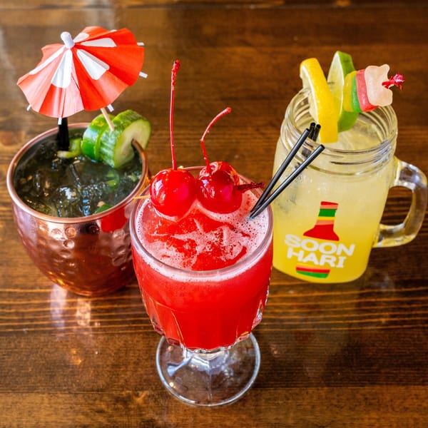 Three colorful Angry Chicken cocktails, garnished with fruit, umbrellas, and candy, on a wooden table.